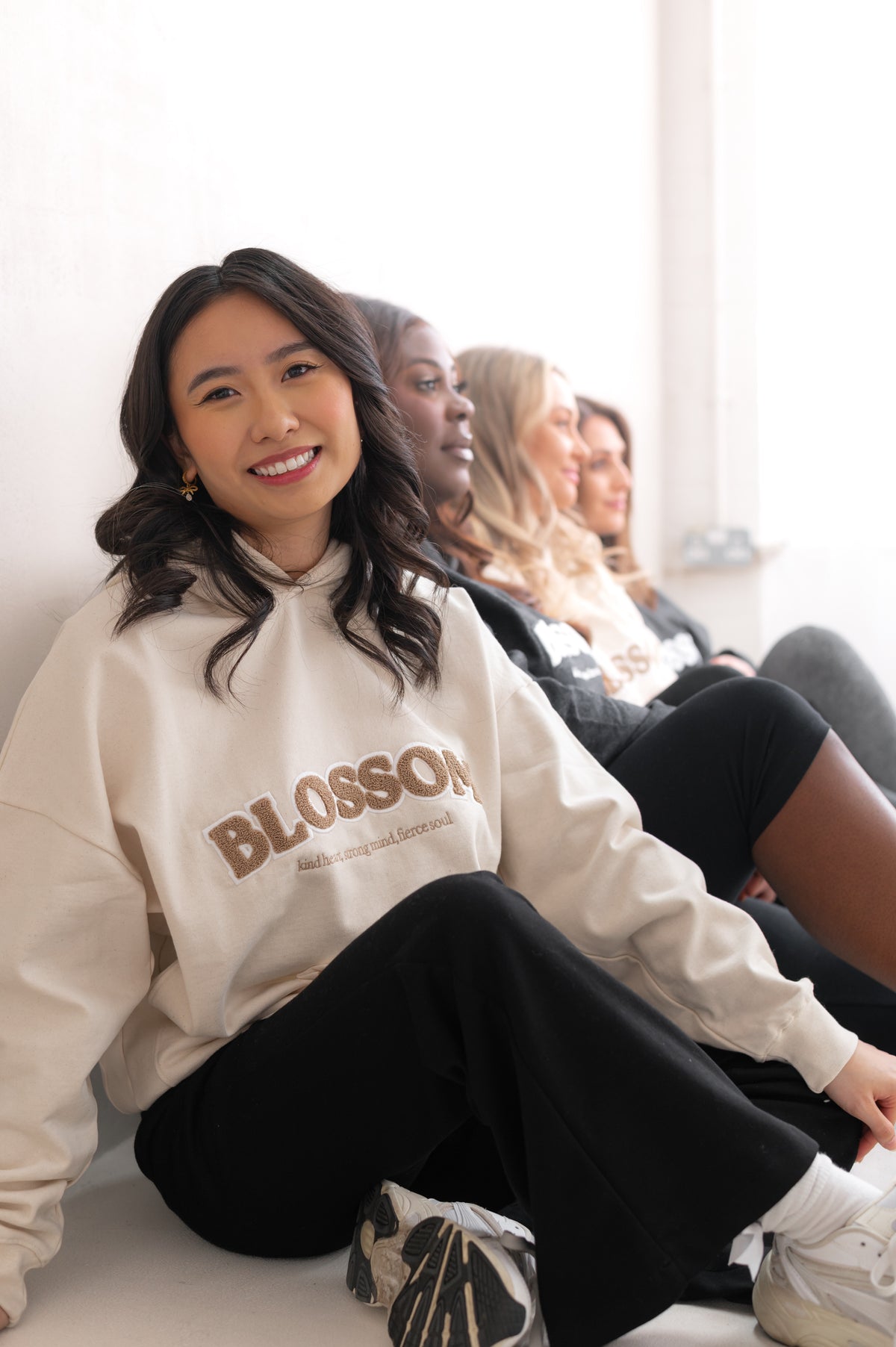 Woman wearing a 'Blossom' sweatshirt sitting on a couch with blurred people in the background wearing the rest of Blossom's original collection.