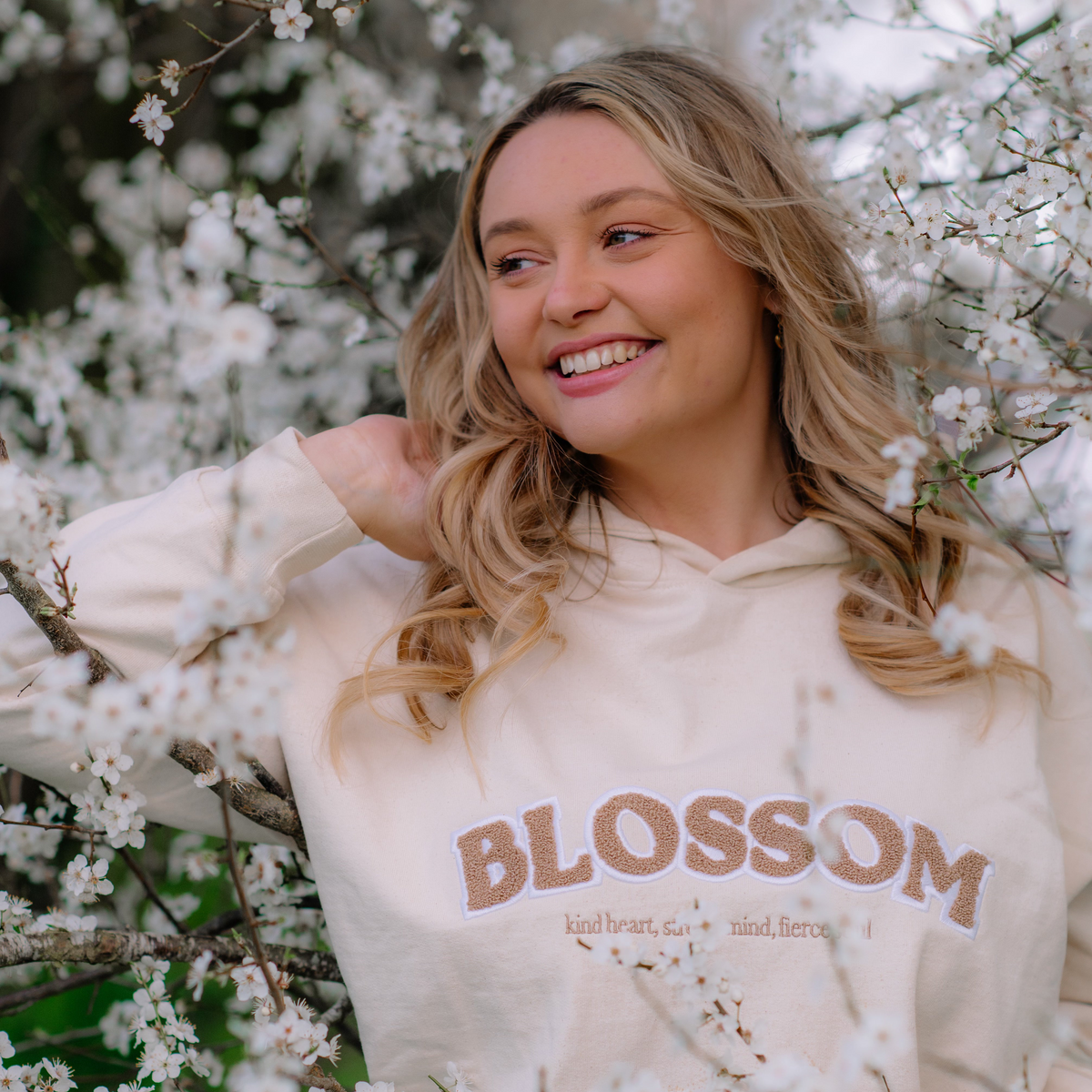 A smiling young woman with blonde hair stands outdoors among white blossoms, wearing a cream hoodie with the word 'BLOSSOM' and smaller text printed across the front that reads 'Kind heart, strong mind, fierce soul'.