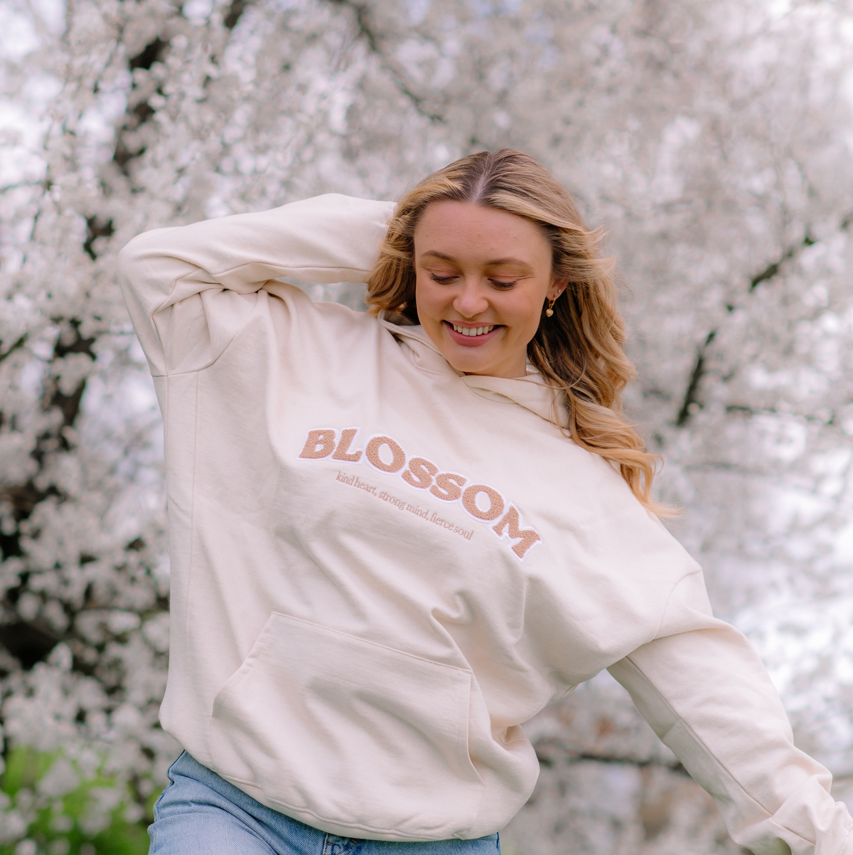 A woman wearing a cream-colored hoodie with 'BLOSSOM' printed in beige across the chest, posing outdoors in front of a background of blooming white cherry blossom trees.