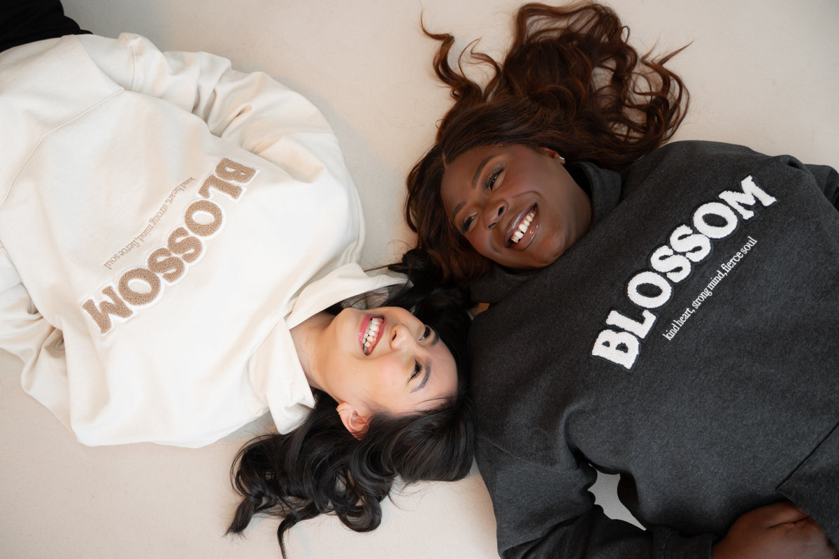 Two women lying down wearing 'Blossom' hoodies and laughing together.