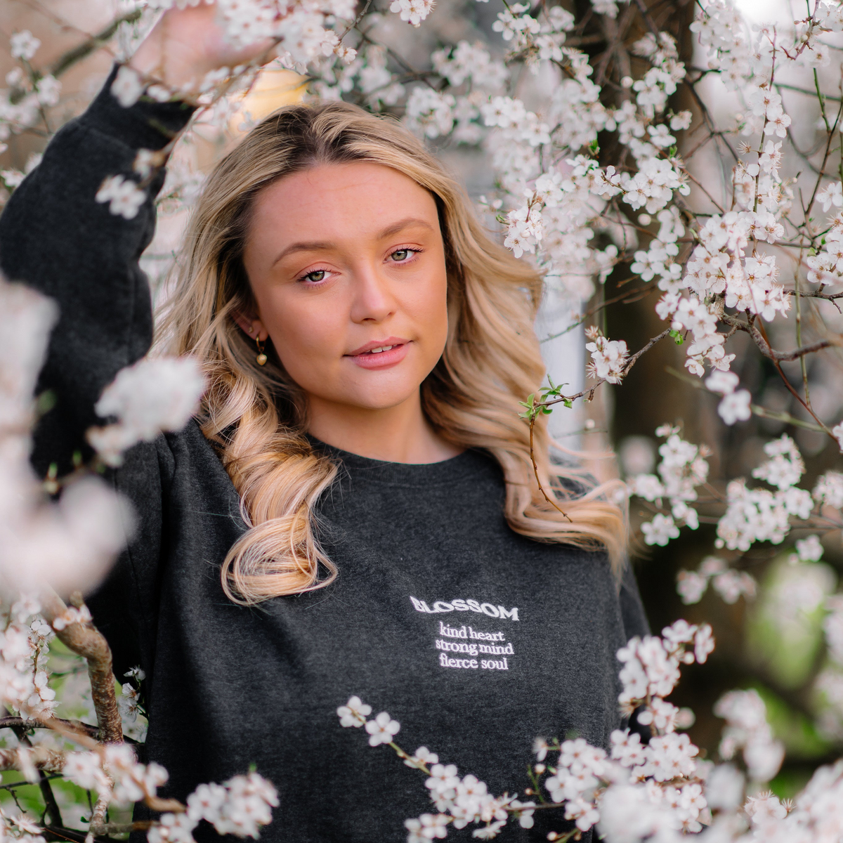 A young blonde woman wearing a dark charcoal gray crewneck sweatshirt with white text that reads 'BLOSSOM kind heart strong mind fierce soul', posing amidst blooming white cherry blossoms.