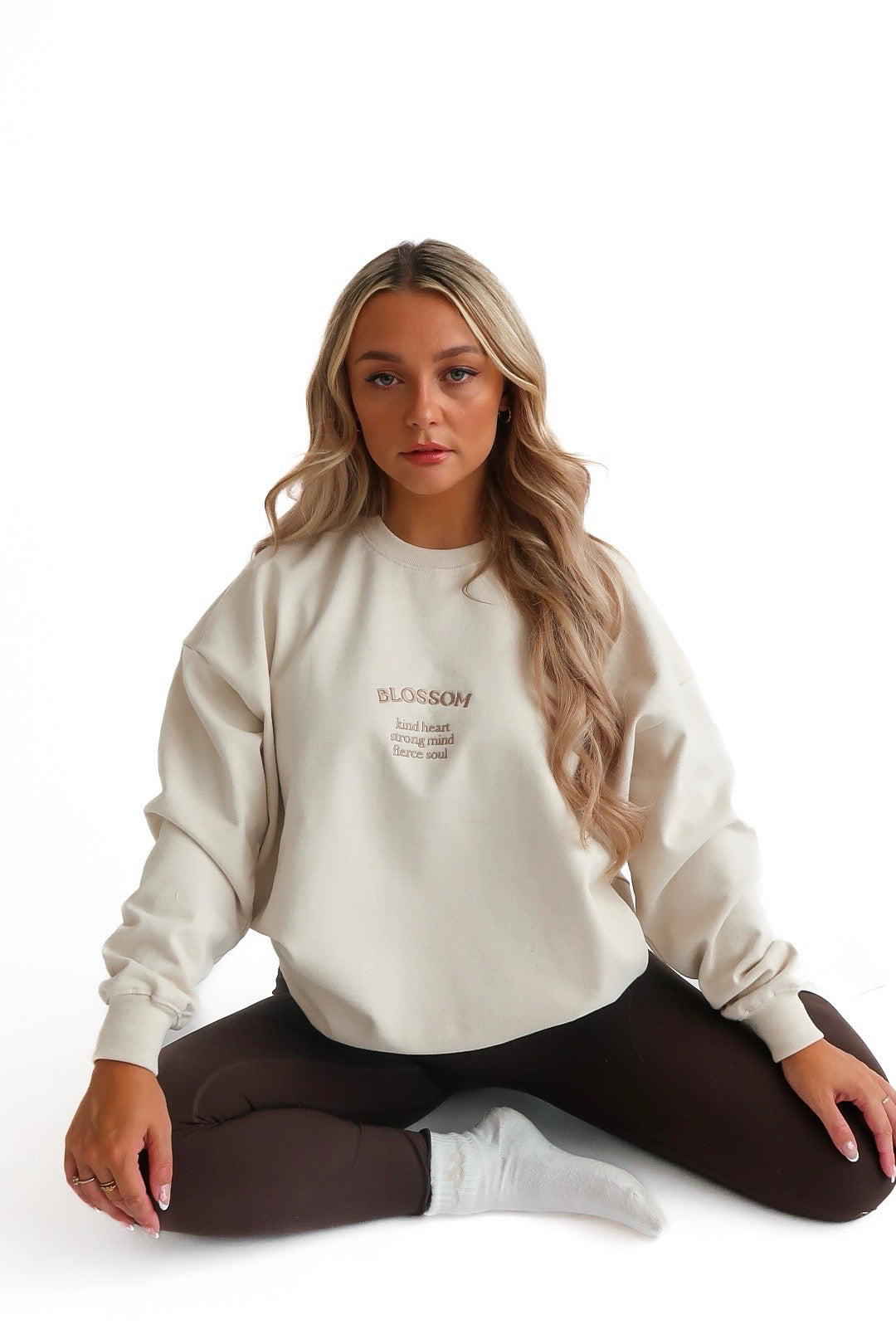 A woman sitting cross-legged wearing a beige cream sweatshirt with the text 'Blossom' on it, paired with dark leggings.