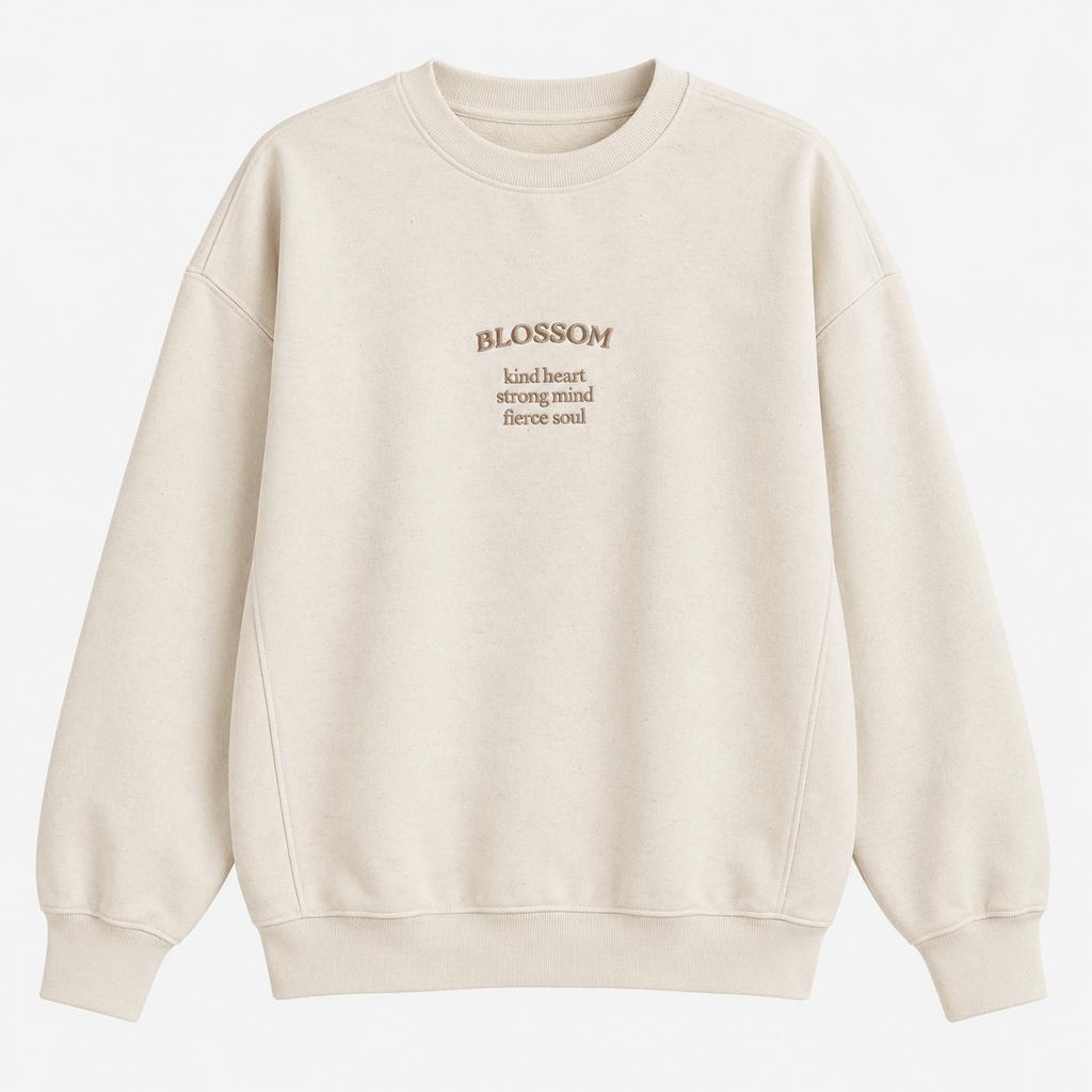 An off-white crewneck sweatshirt with 'BLOSSOM' and three lines of descriptive text 'kind heart, strong mind, fierce soul' printed in a muted brownish-gold tone on the centre chest, displayed on a clean white background.