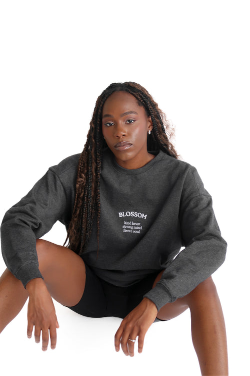 A woman sitting and wearing an oversized gray sweatshirt with the brand name 'Blossom' on it. 