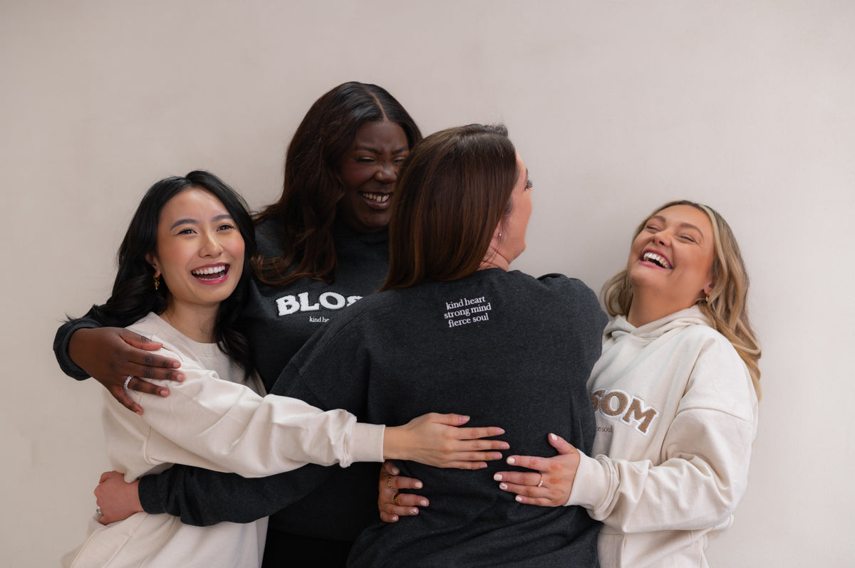 Four people hugging each other against a plain background wearing Blossom The Original Collection. Meaningful hoodies and sweatshirts that read Blossom 'kind heart strong mind fierce soul' designed for empowerment
