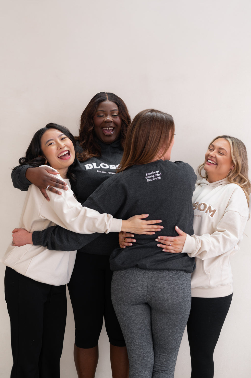 Four women hugging each other against a white background, wearing branded sweatshirts from Blossom with the slogan 'kind heart, strong mind, fierce soul'