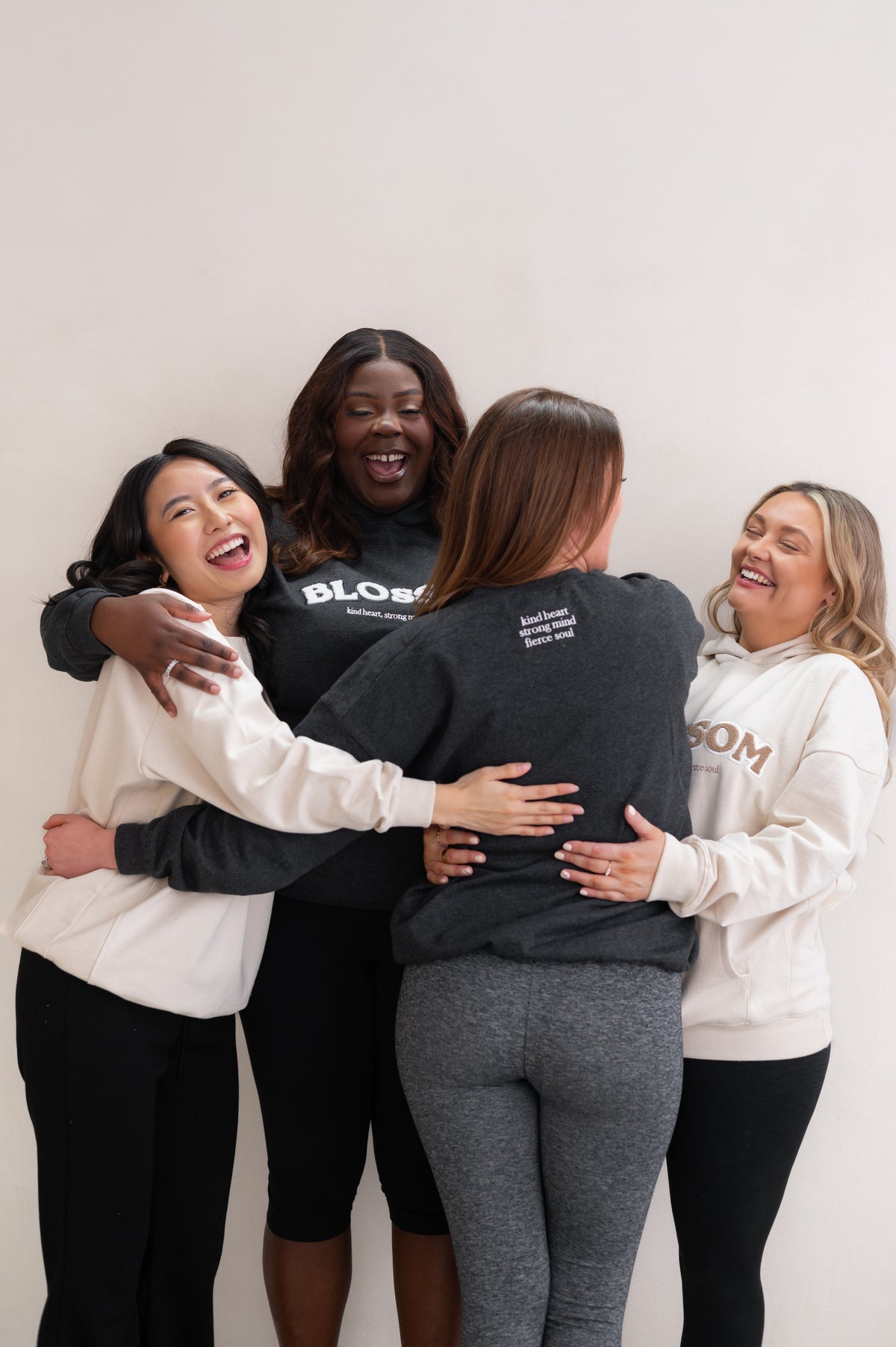 Four women hugging each other against a white background, wearing branded sweatshirts from Blossom with the slogan 'kind heart, strong mind, fierce soul'