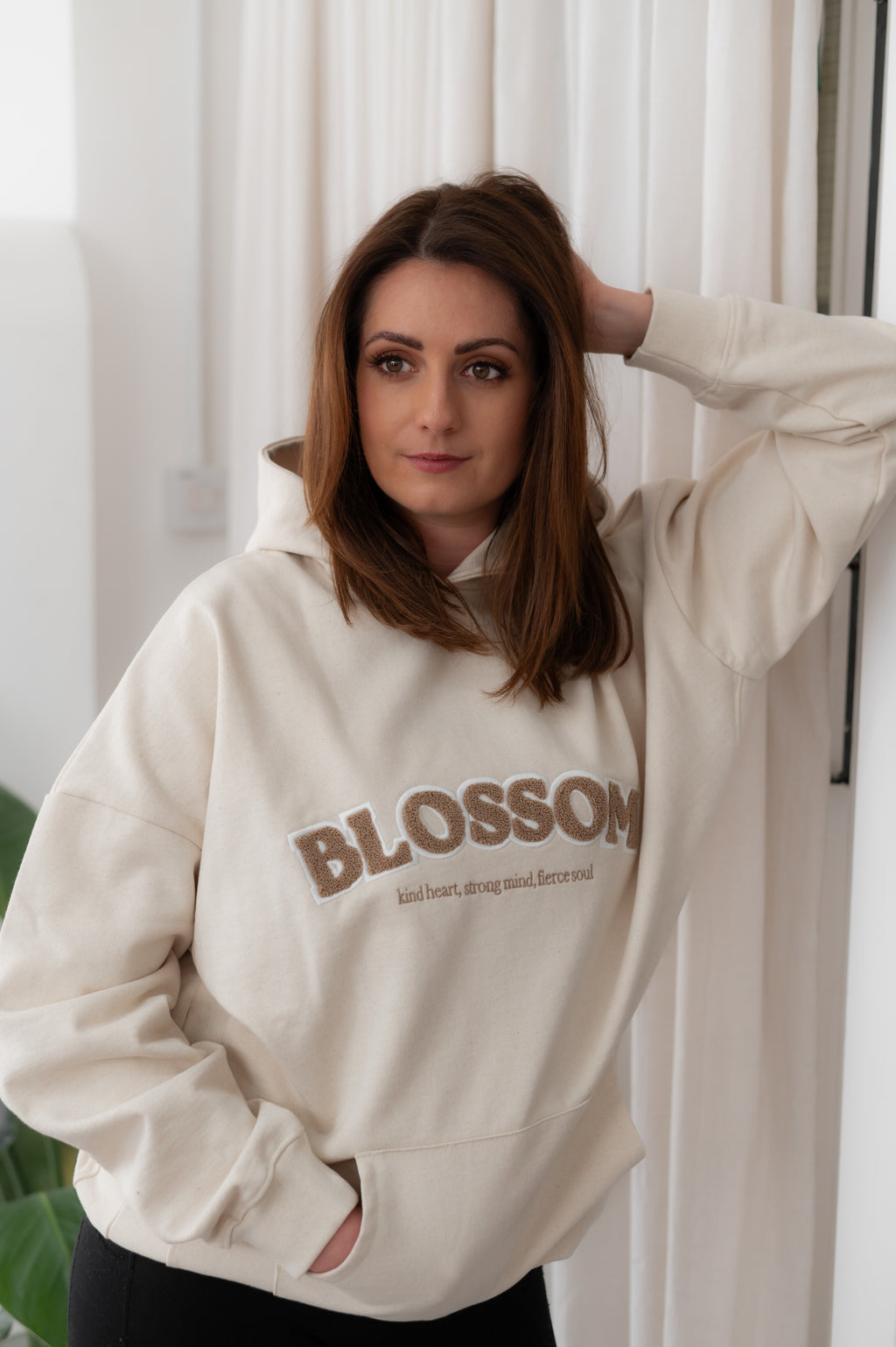 Woman wearing a beige hoodie with 'Blossom' text, standing against a white curtain.