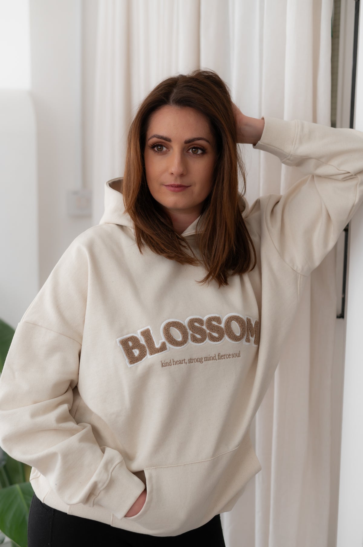 Woman wearing a beige hoodie with 'Blossom' text, standing against a white curtain.