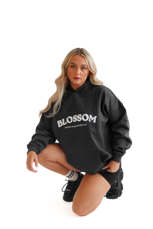 A female model wearing a gray hoodie with the word 'BLOSSOM' printed across the front, posing by crouching down against a plain background.