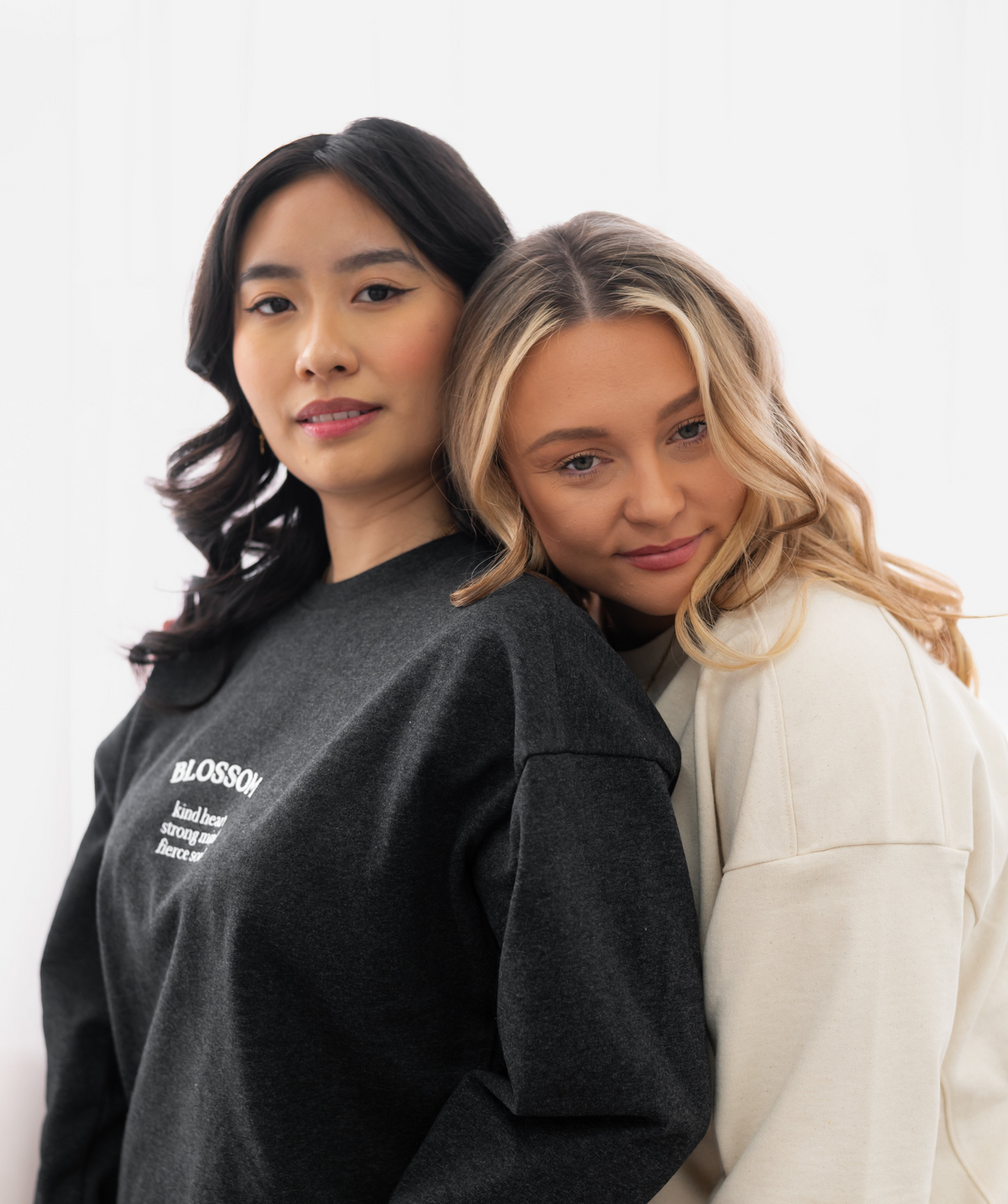 Two women posing together against a clean white background, wearing oversized sweatshirts. The woman on the left wears a dark charcoal gray sweatshirt with white text reading 'GLOSSARY' and a definition, while the woman on the right wears a light cream-colored sweatshirt.